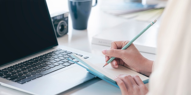 Closeup image of woman hand writing on notebook with pencil while working at her office desk.