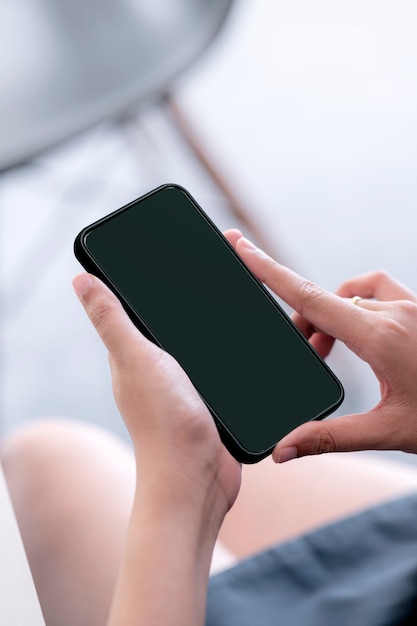 自宅の椅子に座っている間、黒い画面のスマートフォンを保持している女性の手のクローズアップ画像。垂直方向のビュー。