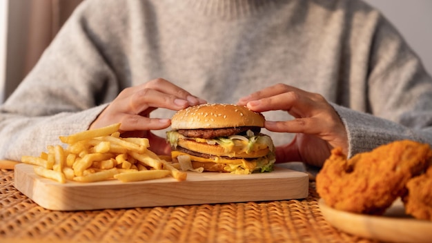 Closeup image of a woman holding and eating hamburger and french fries with fried chicken on the table at home