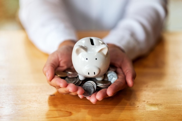 Closeup image of a woman holding piggy bank and coins in hands on the table for saving money and financial concept