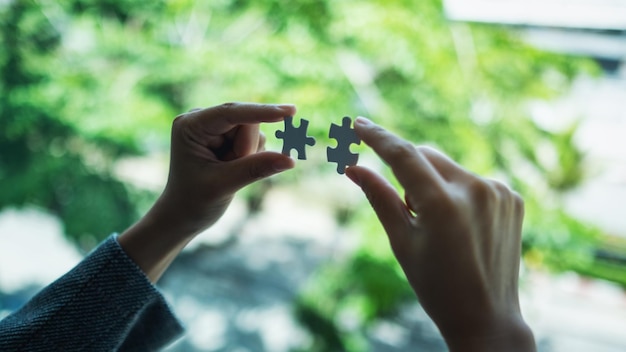 Closeup image of a woman holding and putting a piece of white jigsaw puzzle together with green nature background