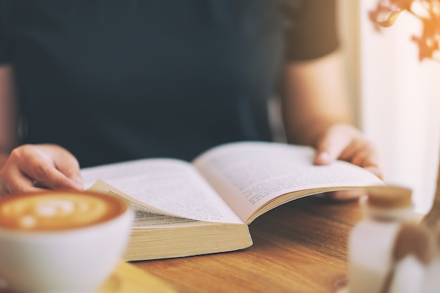 木製のテーブルにコーヒーカップとビンテージ小説の本を持って読んでいる女性のクローズアップ画像
