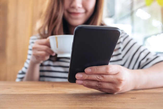 Крупным планом изображение женщины, держащей, использующей и смотрящей на смартфон во время питья кофе