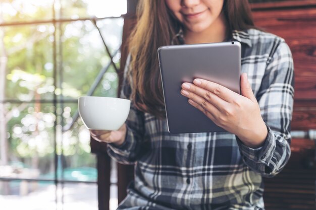 Closeup image of a woman holding and using tablet pc while drinking coffee in cafe