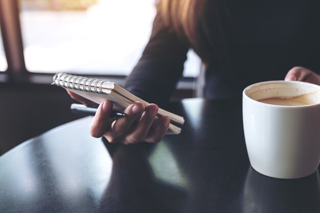 Closeup image of a woman holding a white blank notebook and coffee cup on table 