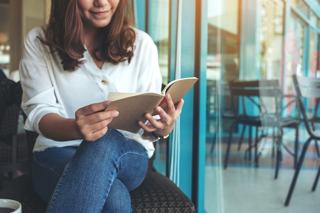 Closeup image of a woman opening a book to read with notebooks and coffee cup on wooden table in cafe