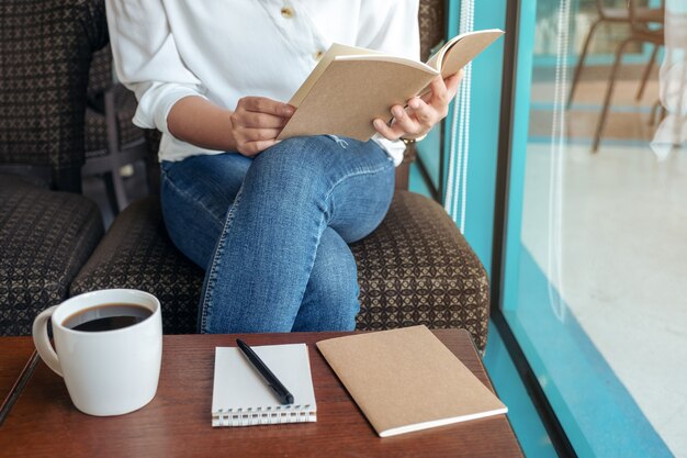 カフェの木製テーブルにノートブックとコーヒーカップで読むために本を開く女性のクローズアップ画像