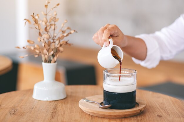 カフェの木製テーブルに氷とミルクのグラスにコーヒーを注ぐ女性のクローズアップ画像