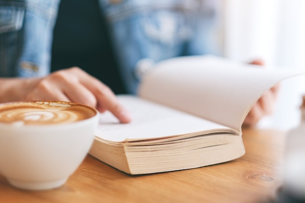Closeup image of a woman reading and pointing at a vintage novel book with coffee cup on wooden table