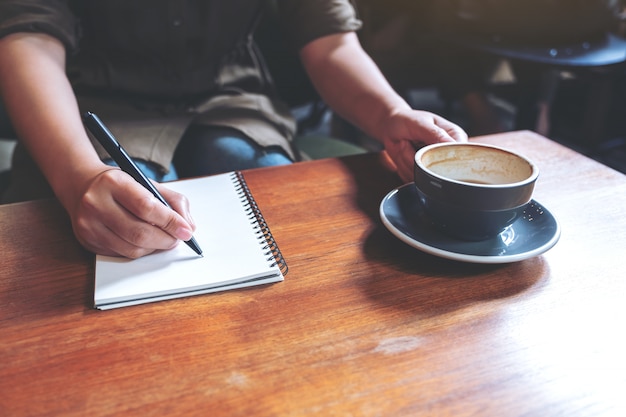 Closeup image of a woman's hand writing down on a white blank notebook while drinking coffee on wooden table