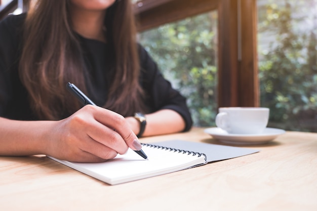 Closeup image of a woman's hand writing down on a white blank notebook with coffee cup on wooden table