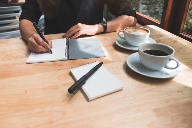 木製のテーブルの上のコーヒーカップと白い空白のノートに書き留めている女性の手のクローズアップ画像