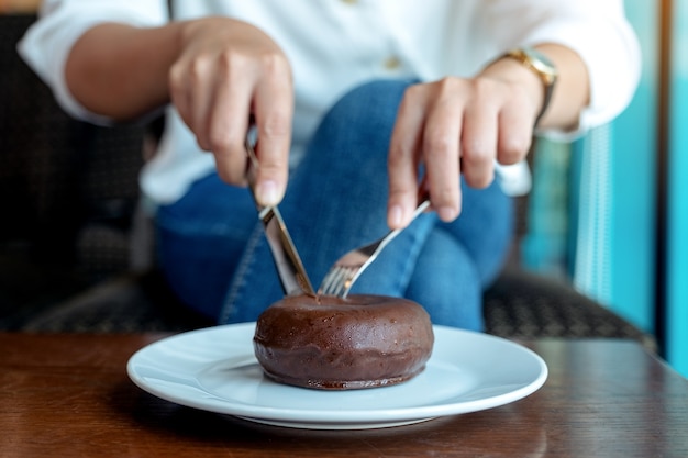 Closeup image of woman's hands cutting a piece of chocolate donut by knife and fork for breakfast on wooden table