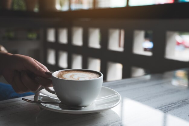 カフェのテーブルでハートカフェラテアートとホットコーヒーのカップを保持している女性の手のクローズアップ画像