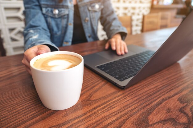 Closeup image of a woman using and touching on laptop touchpad on wooden table while drinking coffee