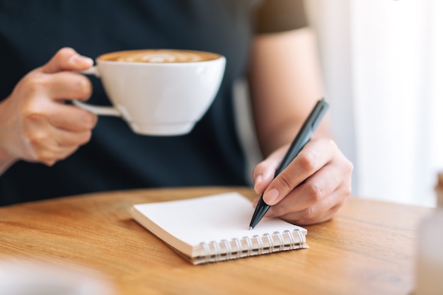 Closeup image of a woman writing on blank notebook while drinking coffee