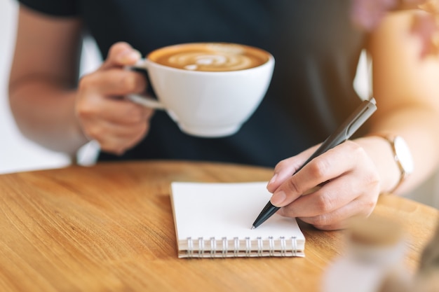 Immagine del primo piano di una donna che scrive su un taccuino bianco mentre beve caffè