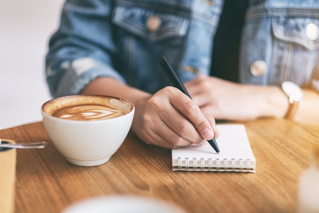 木製のテーブルの上のコーヒーカップと空白のノートに書いている女性のクローズアップ画像