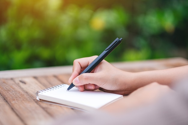 Closeup image of a woman writing on blank notebook on wooden table in the outdoors