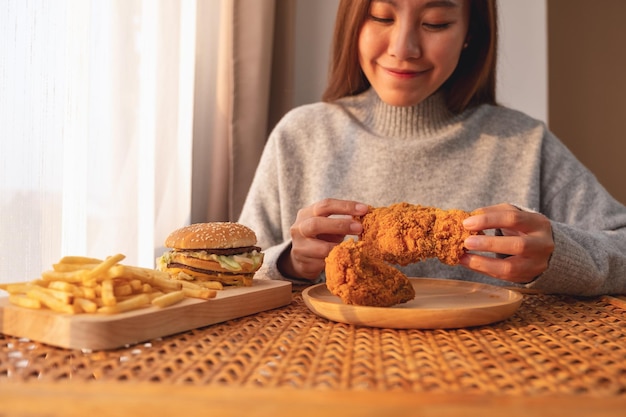 Closeup image of a young asian woman holding and eating fried chicken with hamburger and french fries on the table at home