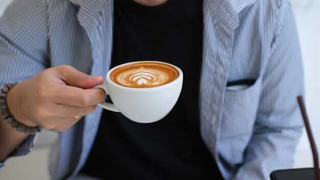 Closeup image of a young man holding a coffee cup with latte art in the comfortable living room