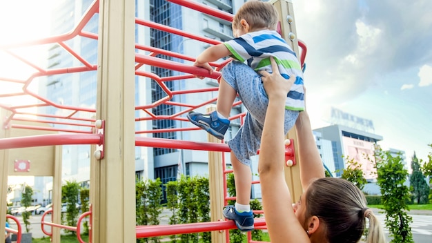 어린 아들이 스포츠 어린이 놀이터에서 높은 금속 계단을 오르는 것을 돕는 젊은 어머니의 근접 촬영 이미지