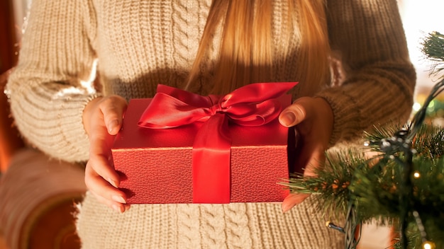 Immagine del primo piano di una giovane donna in maglione che tiene in mano una scatola regalo di natale con un nastro rosso