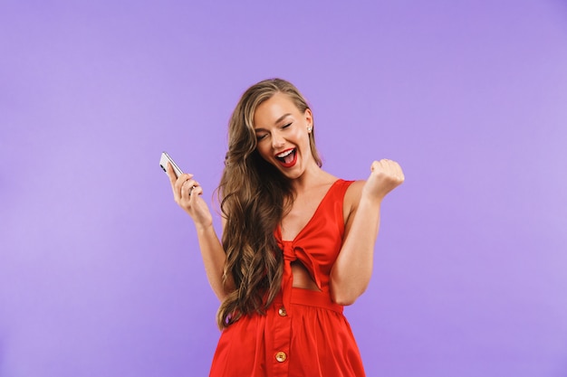 Closeup of joyful young woman wearing red dress screaming and rejoicing while holding mobile phone
