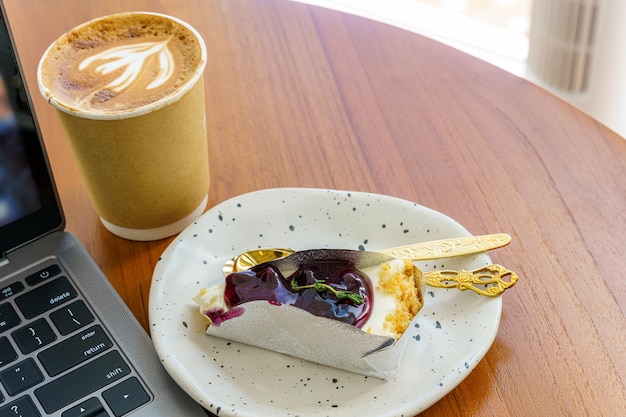 Closeup of keyboard laptop and Hot coffee latte with latte art milk foam in cup made of paper and Blueberry Cake on wood desk on top view In a coffee shop at the cafeduring business work concept