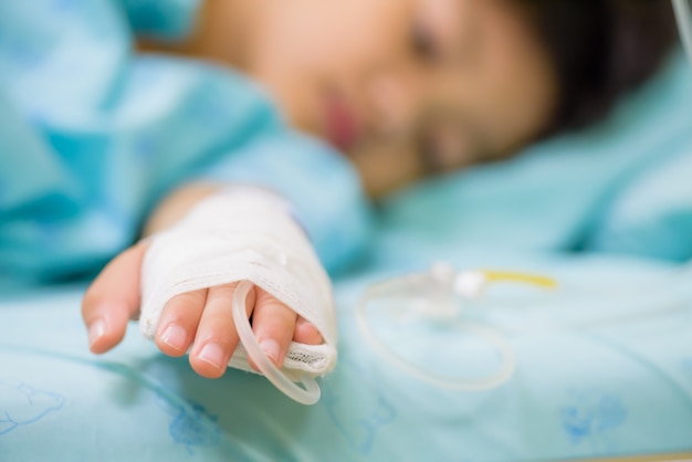 La mano del bambino del primo piano dorme su un letto in ospedale con endovenoso salino, fuoco selettivo.