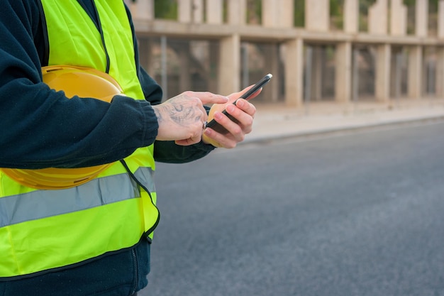 Closeup of a laborer looking at a phone while holding a yellow hard hat with a construction site in the background