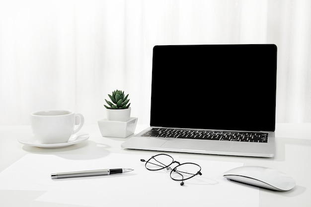 Closeup of a laptop, a cup of coffee, glasses, and more on a white desk, indoors