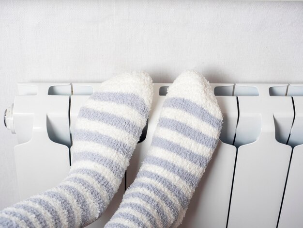 Closeup of legs dressed in warm socks on a white central heating battery The concept of the winter season the heating crisis High gas prices