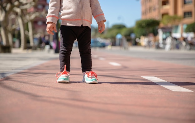 Крупным планом ноги маленькой девочки в кроссовках и черных леггинсах, стоящих над взлетно-посадочной полосой в солнечный день