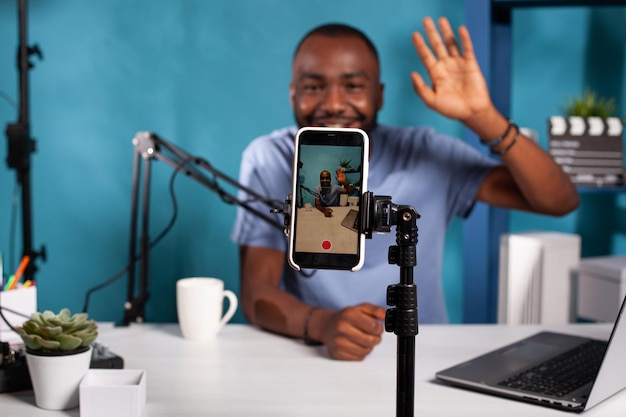 Closeup of live vlog setup with smarthone on stand filming\
vlogger waving hello sitting at desk with professional microphone.\
selective focus on mobile phone recording social media\
influencer.