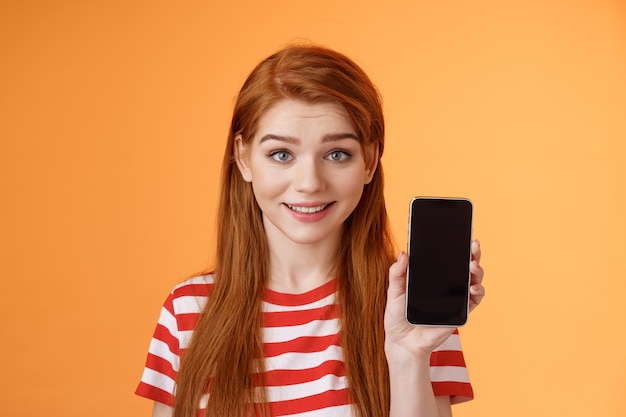 Closeup lovely caucasian redhead girl showing smartphone screen orange background smiling cute recom...