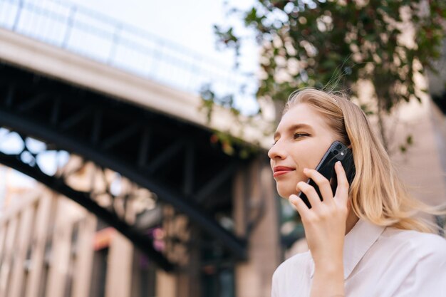 Closeup lowangle view face of attractive blonde lady talking on mobile phone holding smartphone close to ear looking away sanding alone on city street