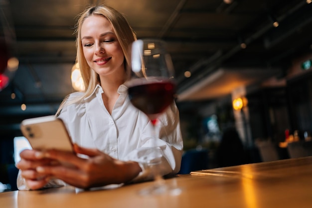 Closeup lowangle view of smiling blonde young woman using mobile phone typing online message sitting at table with glass of red wine at restaurant