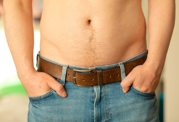 Foto primo piano di una pancia maschio. un uomo mostra la pancia nuda con le mani nelle tasche dei pantaloni jeans