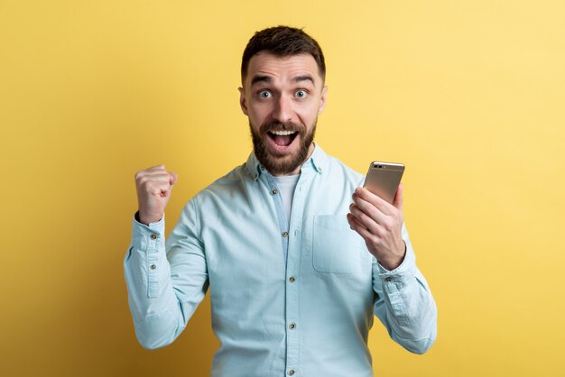 Fronte maschio del primo piano che tiene uno smartphone e sguardo emozionante che serra un pugno nella posa di vittoria. vincitore dell'emozione