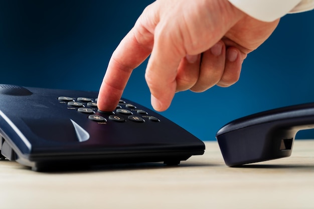 Closeup of male finger pressing a button on landline telephone keypad on a desk. Over dark blue background.