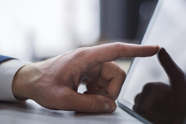 光のぼかしオフィスの背景に机の上の現代のデジタル タブレットの画面に触れる男性の指のクローズ アップ