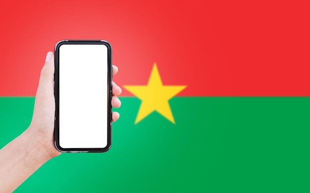 Primo piano della mano maschile che tiene smartphone con schermo vuoto sullo sfondo della bandiera sfocata del burkina faso