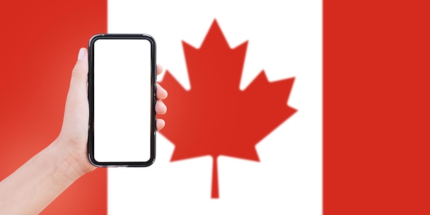 カナダのぼやけた旗の背景に画面に空白のスマート フォンを持っている男性の手のクローズ アップ