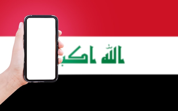 Primo piano della mano maschile che tiene lo smartphone con lo schermo in bianco sullo sfondo della bandiera sfocata dell'iraq