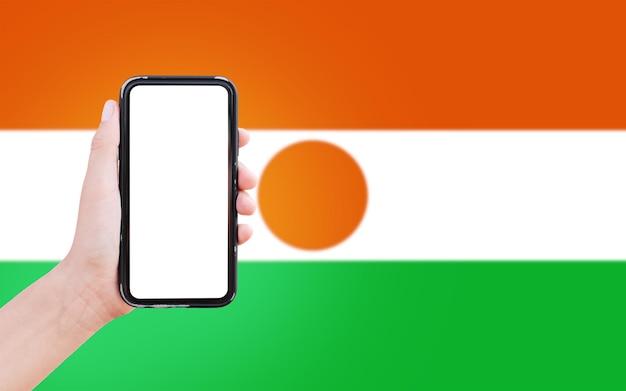 Closeup of male hand holding smartphone with blank on screen on\
background of blurred flag of niger