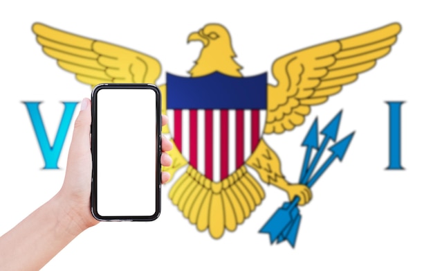 Closeup of male hand holding smartphone with blank on screen on background of blurred flag of United States Virgin Islands