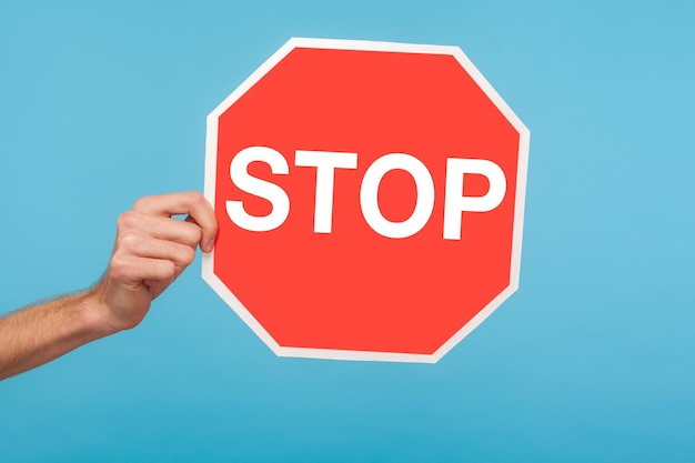 Photo closeup of male hand holding stop symbol showing red traffic sign warning about problems concept of ban denial prohibited to go forbidden way indoor studio shot isolated on blue background