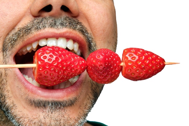 Closeup of male mouth with short beard biting a skewer with three strawberries on pure white background