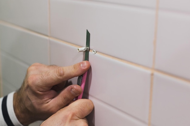 Closeup of man hands cutting extra part of dowels hammered into tiled wall in washroom with stationery knife Renovation works in bathroom Dirt from renovation process